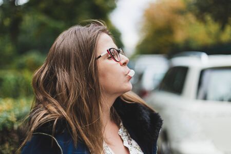 Beautiful girl making chewing gum bubbles outdoor - Cute young woman with brown hair wearing eyeglasses holding white gum in her mouth - Pretty female having fun in the cityの写真素材