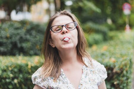 Young woman chewing gum and making small bubble outdoors - Fashionable cute girl wearing eyeglasses...