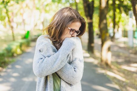 Young woman trembling and feeling cold outdoors - Thick dressed girl with grey jumper wearing eyeglasses freezing while walking in the park in autumnの写真素材
