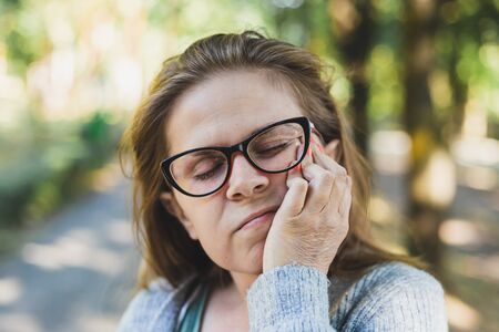 Young woman wearing eyeglasses having a dental pain while looking down outdoors - Cute girl holding one hand on her face while suffering from a toothacheの写真素材