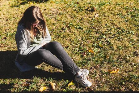 Young woman with menstrual pain sitting on the grass on a sunny day - Casually dressed girl with brown hair suffering form cramps and discomfort in the abdominal area caused by the periodの写真素材