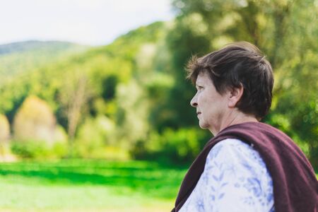 Beautiful senior woman standing alone in nature on a summer day - Sad old lady feeling lonely and looking in the distance while walking in the forestの写真素材