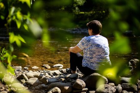 Senior woman sitting alone and admiring a river in the forest - Dramatic concept of an old lady...