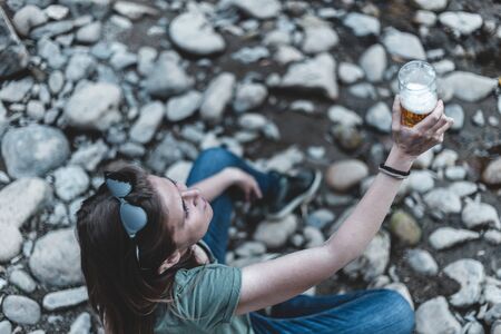 Young woman holding a glass of fresh beer while standing near a river - Pretty girl cheering with a pint of cold beverage in nature - Happy casually dressed female rising mug of alcoholic drinkの写真素材