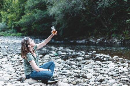 Happy young woman cheering with a glass of beer while sitting by a river in the forest - Casually dressed attractive girl rising a pint of fizzy and cold beverage in nature on a summer dayの写真素材