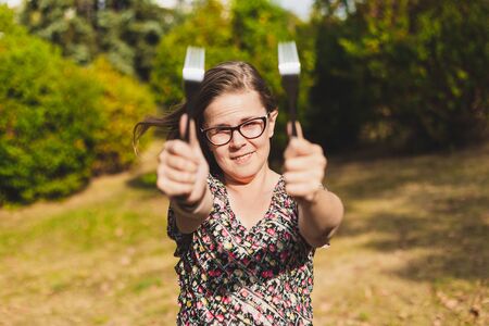 Happy woman wearing eyeglasses having fun while playing with two forks outdoors - Casually dressed smiling girl with brown hair feeling good and acting silly in the parkの写真素材