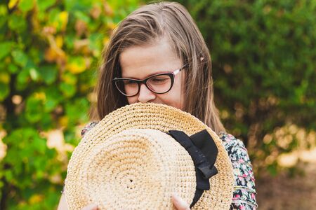 Shy young woman standing in the park while holding summer hat against her face - Casually dressed...