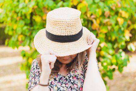 Shy woman covering her face with a summer hat outdoors - Pretty girl feeling humiliated while hiding behind straw hat in the park - Cute and adorable gesture made by young female in sign of shameの写真素材
