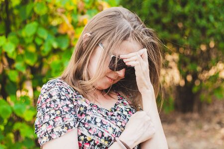 Smiling shy young woman looking down while holding one hand to her forehead outdoors - Cute girl with brown hair wearing sunglasses feeling embarrassed and trying to cover her face in the parkの写真素材