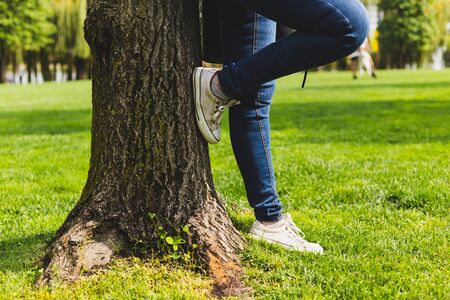 Person wearing sneakers and jeans while leaning against a tree in the park - Casually dressed millennial girl with canvas shoes and denim pants standing on the grass outdoorsの写真素材