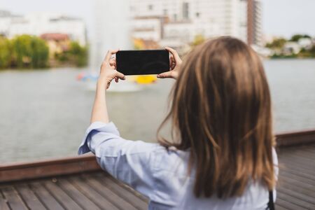 Rear view of young woman taking photos to lake landscape with the mobile phone - Pretty girl with brown hair and blue shirt making nature pictures - Relaxing and fun activityの写真素材