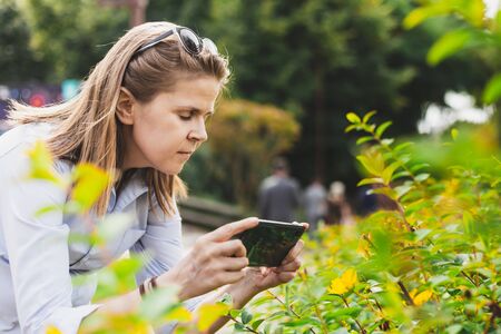 Pretty woman taking photos of flowers with small mobile phone in the city - Girl with brown hair wearing white shirt having fun in the city while shooting images of floral decorations with phoneの写真素材