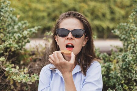 Young girl eating a biscuit outdoors - Casually dressed office woman in a shirt wearing sunglasses having a quick snack in a public parkの写真素材