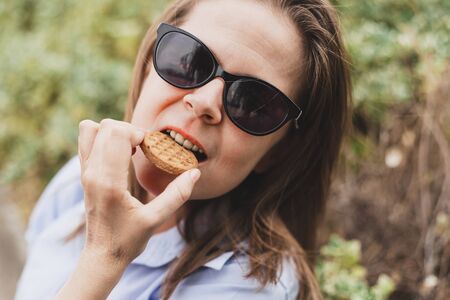 Young woman wearing sunglasses eating a cereal biscuit outdoors - Portrait of a beautiful girl with...
