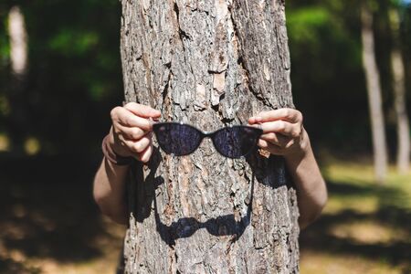 Young woman holding black sunglasses in front of a thin tree trunk in the forest - Girl having fun...