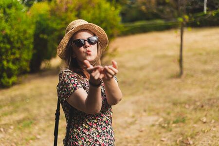 Beautiful young woman sending air kiss outdoors - Casually dressed millennial wearing summer hat and sunglasses sharing love while walking in the park - Adorable girl showing affectionate gesture in natureの写真素材