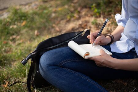 Woman taking notes in a small notebook while sitting down in a park - Casually dressed girl making calculations and analysis outdoorsの写真素材