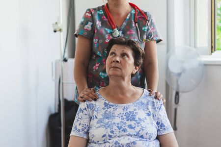 Nurse holding senior woman while sitting on a chair in a hospital - Female medical assistant with pretty old lady in a private clinic - Concept image for elderly health careの写真素材