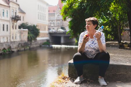 Happy senior woman holding water bottle and red apple while sitting outdoors on a summer day - Casually dressed cute old lady whit healthy diet relaxing in the city on a bright sunny dayの写真素材