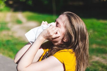 Sick young woman having a cold blowing her nose outside - Pretty girl with brown hair suffering from summer allergies in the parkの写真素材