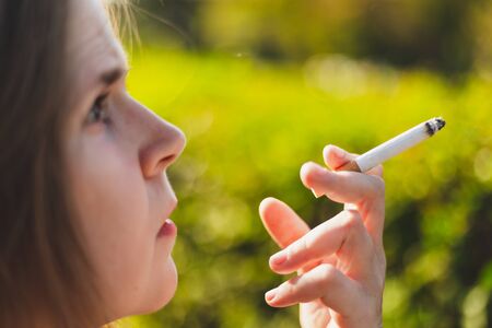 Close-up shot of young woman smoking outside - Pretty girl with dangerous and toxic habit holding a lit cigar in the parkの写真素材
