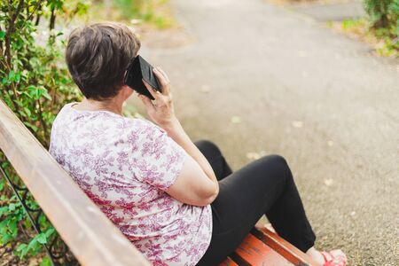 Rear view of senior woman talking on the phone while sitting on a bench in the park - Old lady using modern cell phone for socializing and communicating with others - Concept image for elderly people using technologyの写真素材