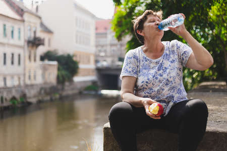 Senior woman drinking from a water bottle for hydration in the heat while sitting in the sun and holding an halved appleの写真素材