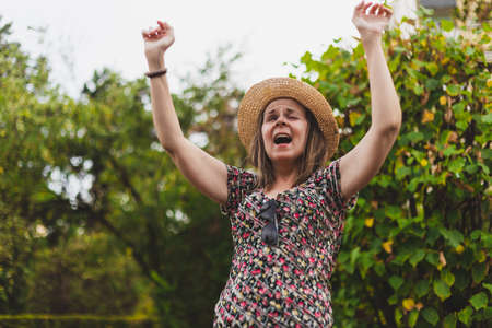 Cute young woman screaming while holding both hands in the air outside - Casually dressed teen girl wearing straw summer hat and floral shirt yelling to someone while lifting arms up in the parkの写真素材