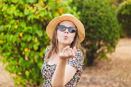 Beautiful happy millennial girl sending air kiss outdoors - Pretty smiling young woman wearing summer straw hat and sunglasses sharing love and affection in the parkの写真素材