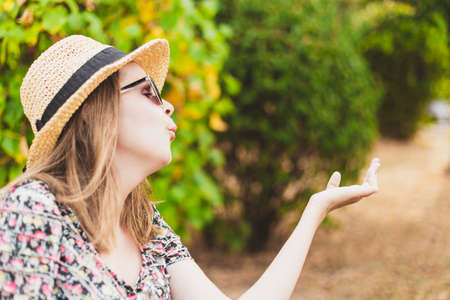 Beautiful young woman blowing air kiss with copy space on a summer day in nature - Hipster girl wearing straw hat and summer glasses sharing love and affection while feeling good in the park on a bright sunny dayの写真素材