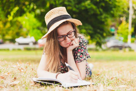 Pretty girl writing a book outside - Attractive person with eyeglasses and straw hat taking notes into her notebookの写真素材