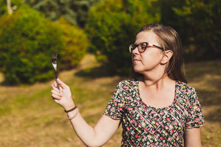 Beautiful young woman wearing eyeglasses holding a fork outside - Hungry cute teen girl acting silly and feeling good in nature - Playful female with kitchen utensil in the parkの写真素材