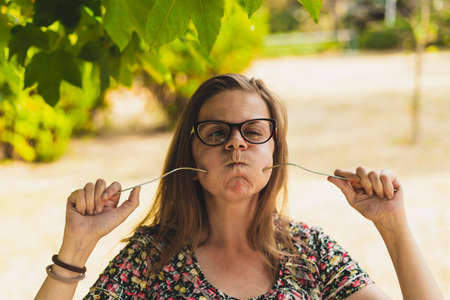 Cute girl having fun playing with a fork outdoors on a sunny day - Pretty young woman with brown hair being funny and acting crazy while sticking kitchen utensil into her cheek in the parkの写真素材