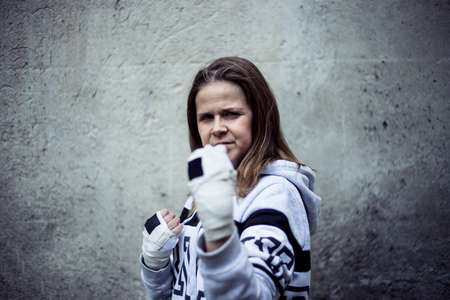 Smiling young female fighter taking defense position in an urban scenery - Strong and confident girl with brown hair wearing white hoodie standing in a black position - Concept image for woman powerの写真素材