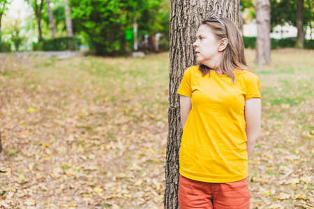 Beautiful young woman hiding scared behind a tree in the park on a summer day - Casually dressed teen girl with brown hair suffering from anxiety while sitting alone in nature - Having a panic attackの写真素材