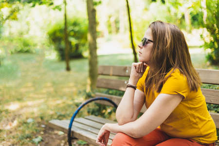 Thinking woman looking forward - Thoughtful girl sitting on bench - Lonely girl - Teenager waiting outside while daydreamingの写真素材