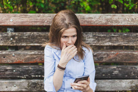 Beautiful young woman looking scared and surprised while sitting on a bench in the park on a summer day - Pretty girl with brown hair wearing a casual outfit standing alone while having a panic attack outdoorsの写真素材