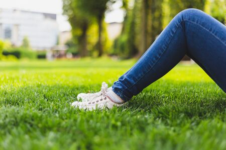 Close-up shot of woman wearing canvas shoes and jeans sitting on the green grass in the park - Casually dressed teen girl with white trendy sneakers - Modern and urban woman foot wear in nature on a summer dayの写真素材