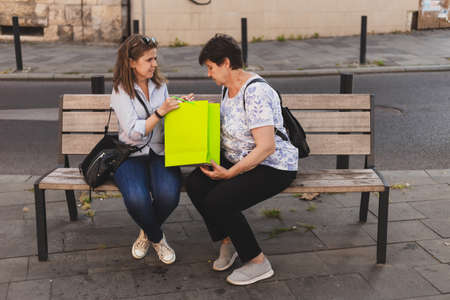 Happy mother and daughter sitting on a bench while looking in colorful shopping bag in the city - Pretty girl and cute old lady spending quality time together outside - Family doing fun and relaxing activityの写真素材