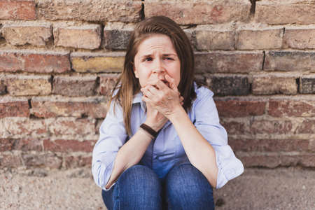 Scared woman sitting on the ground while leaning with elbows to her knees against a brick wall and looking to the camera - Frightened young girl holding her hands to the mouthの写真素材
