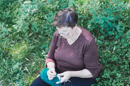 Cute old lady knitting outside in a summer day - Beautiful old lady making winter clothes while sitting in the grass in nature - Elderly person with relaxing hobbyの写真素材