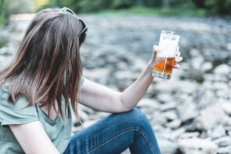 Rear view of young woman cheering with a glass of cold beer in nature near a river â Pretty girl with brown hair relaxing outdoors with a cold summer beverageの写真素材