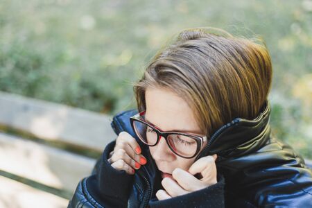 Beautiful woman with eyeglasses freezing outside - Pretty girl with brown hair wearing a bulky jacket feeling cold while sitting in the parkの写真素材