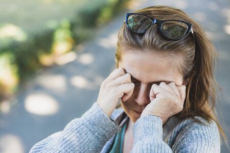Stressed woman with headache rubbing her eyes outside - Tired cute girl with eye pain feeling anxiety symptoms in the park - Concept image for stress managementの写真素材