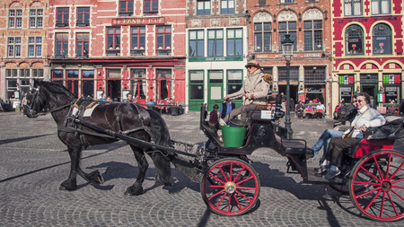 BRUGES, BELGIUM - MARCH 8, 2014: Tourists riding a Horse carriage in front of famous old colorful buildings at Market square. Popular Flemish city with almost intact medieval architecture.のeditorial素材