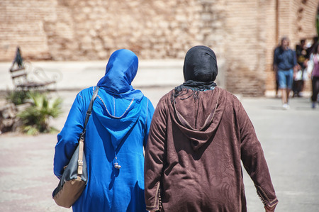 Two moroccan women seen from the back dressed in typical blue and brown djellaba walk down the streets of Marrakesh Medina, Moroccoの写真素材