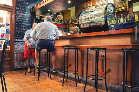 MADRID, SPAIN - JULY 23, 2015: Two local senior men sitting inside a typical spanish tapas bar. Menu written with white chalk on a black board. Different food and drinksのeditorial素材