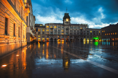 Unity of Italy Square in Trieste, Italy at night during the heavy raining. Illuminated buildings - town hall and cloudy sky. People with umbrellasの写真素材