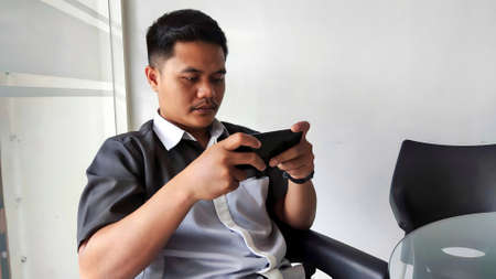 An authorized Indonesian car dealer salesman is waiting for customers to come while playing games on his android smartphoneの写真素材