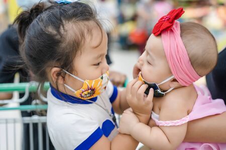 Closeup of an adorable Asian little girl is wearing for younger sister, a beautiful mask against corona virus at the shopping mall. Coronavirus and pollution protection for healthcare concept.の写真素材